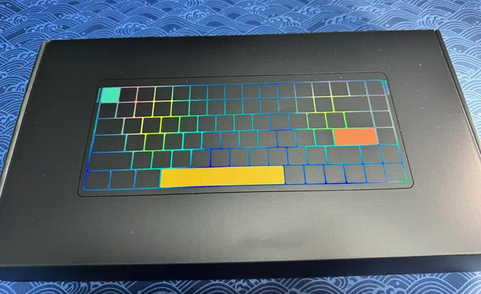 L'image montre l'emballage noir du clavier avec une silhouette de touches aux reflets holographiques multicolores bleus et oranges, posé sur un fond bleu à motifs vagues.