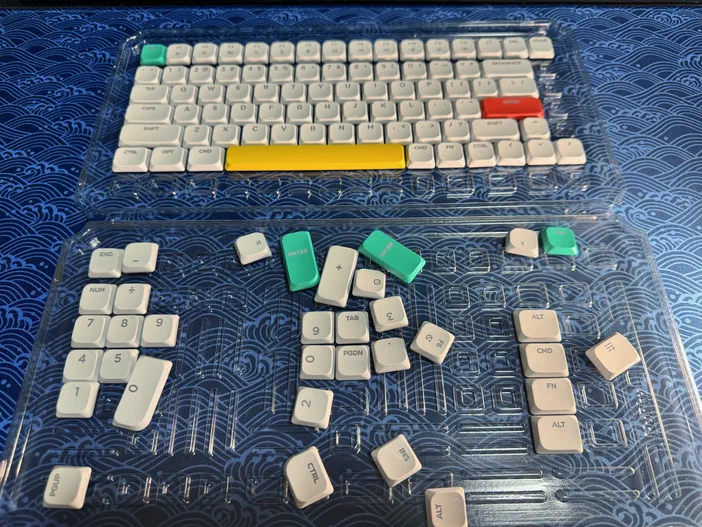 Deux plateaux transparents contenant des keycaps blancs et colorés d'un clavier Nuphy Air75 V2, le premier rempli et le second partiellement vide sur fond bleu.