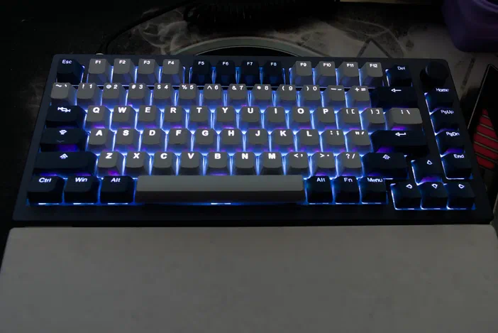 Clavier mécanique compact avec des touches gris foncé illuminées par un éclairage RGB bleu vif sur fond sombre.