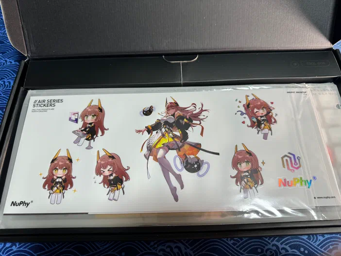 Une planche d’autocollants illustrée par des personnages manga aux cheveux bruns et cornes jaunes, protégés par du plastique transparent à l’intérieur d’un boîtier noir portant le logo NuPhy.