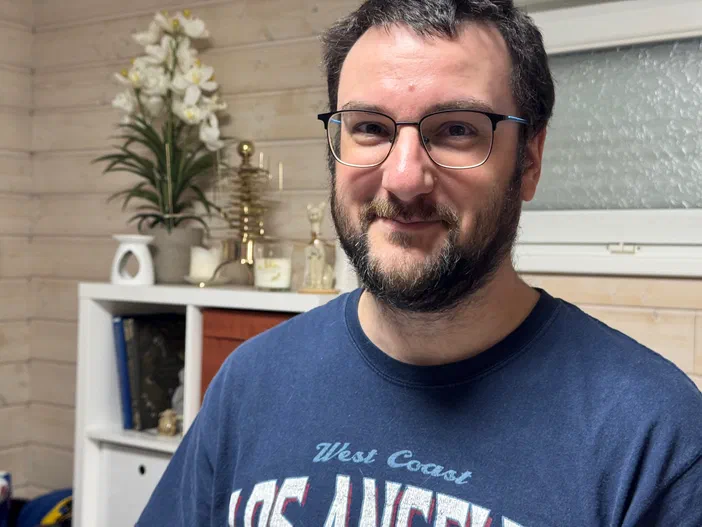 Portrait d'un homme à lunettes et barbe, souriant devant une étagère décorée de fleurs blanches.
