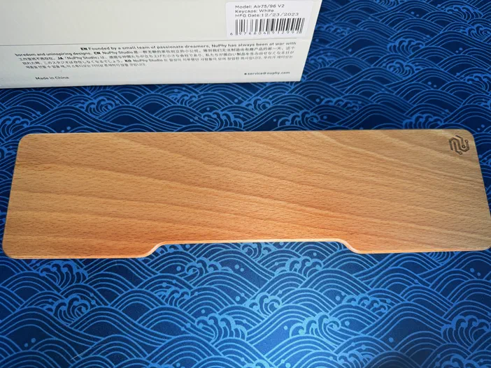 Repose-poignet en bois clair avec logo gravé, posé sur un tapis de bureau bleu à motifs ondulés.