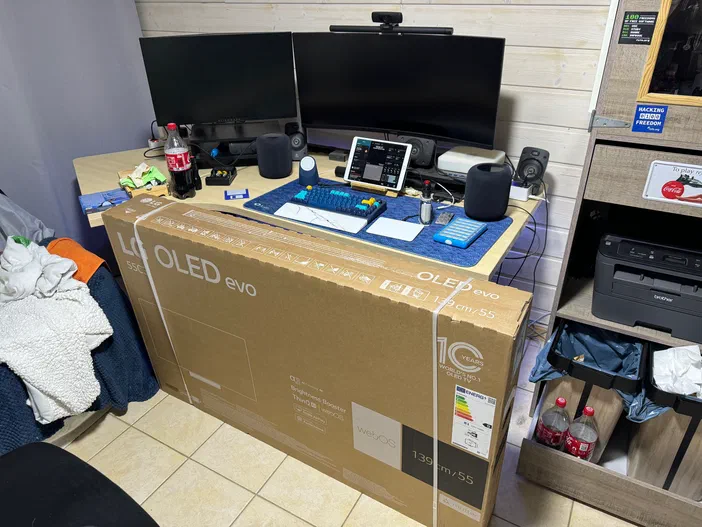 Bureau informatique avec deux écrans, un grand carton d'écran OLED devant et une imprimante sur étagère.