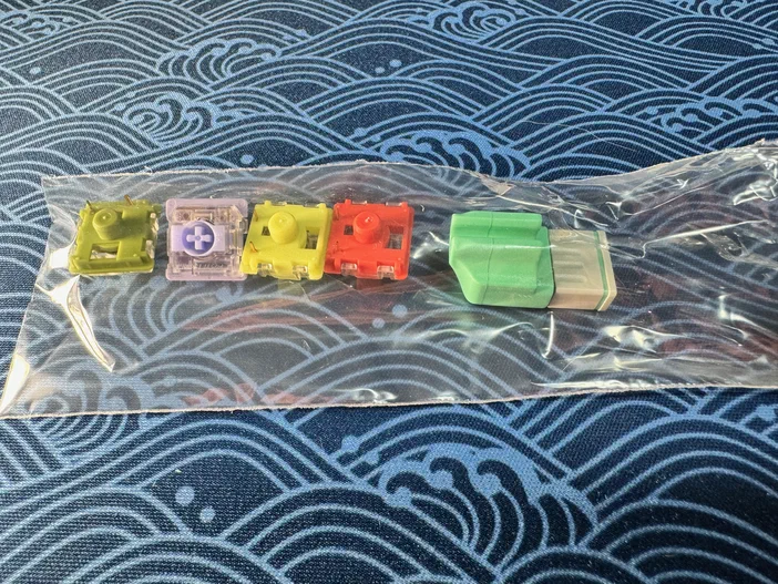 Un sachet transparent contenant cinq switches de clavier colorés (jaune, violet, jaune vif, orange) et un dongle sans-fil vert.