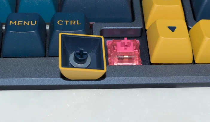 Vue rapprochée d'un clavier personnalisé avec des touches bleues et jaunes ; une touche fléchée jaune est retirée, laissant un anneau de stabilisateur gris visible dans le keycap.